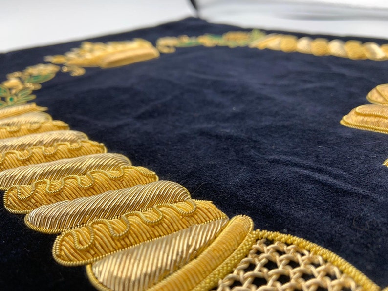 May include: A black velvet cushion with gold embroidery. The embroidery features a crown, a chain, and other decorative elements.