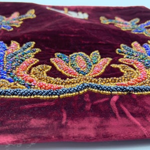 May include: A close-up of a burgundy velvet pillow with a beaded floral design. The design features a variety of colors, including blue, red, gold, and black.