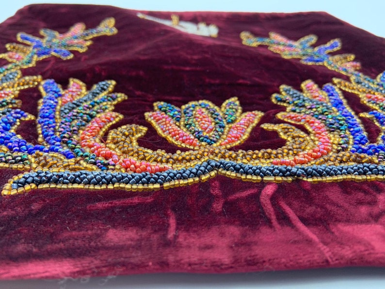 May include: A close-up of a burgundy velvet pillow with a beaded floral design. The design features a variety of colors, including blue, red, gold, and black.