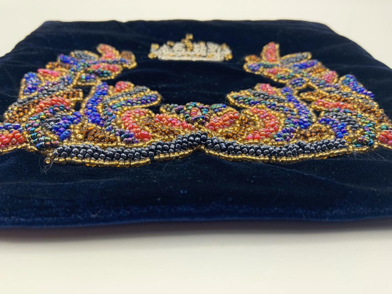 May include: A black velvet pouch with a beaded design of a crown surrounded by leaves. The beads are in shades of red, blue, gold, and iridescent.