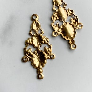 May include: Two gold-toned metal filigree connectors featuring an elongated, symmetrical floral design. Each piece has a central flower motif with radiating petals and small circular loops at the top, bottom, and sides for attachment purposes.