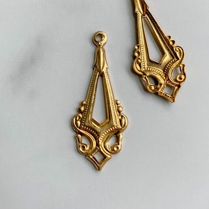 May include: Two gold-toned metal jewellery charms featuring an elongated, open-work diamond shape with intricate scrollwork details and a small circular loop at the top for attachment. The pieces have a polished finish and symmetrical patterns.