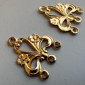 May include: Two gold-toned metal jewelry connectors, each featuring a symmetrical, openwork design with small floral accents at the top and bottom. Each piece includes four small circular loops for attaching chains or other jewelry components.