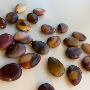 May include: A collection of teardrop-shaped beads in various shades of brown, cream, and burgundy. Each bead has a hole for stringing, suggesting they are for jewelry making or crafts. The beads are smooth and polished.