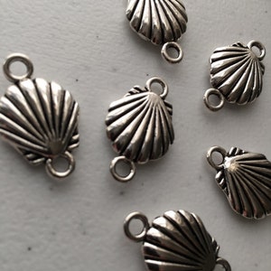 May include: A collection of six silver-toned metal shell charms, each featuring a fan-shaped design with radiating ridges and two small circular loops positioned at the top and bottom for attachment in jewellery making or craft projects.