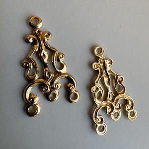 Peut inclure: Deux connecteurs de lustre en métal doré présentant un design ornemental à volutes ajourées. Chaque pièce possède une boucle circulaire unique en haut pour la fixation et trois boucles circulaires plus petites en bas, avec une finition polie.