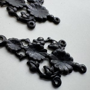 Może przedstawiać: Dwa ciemnoszare metalowe łączniki filigranowe z motywem kwiatowym przypominającym stokrotki i liście. Każdy element posiada cztery okrągłe oczka na końcach i po bokach, przeznaczone do łączenia z innymi komponentami w projektach biżuterii.