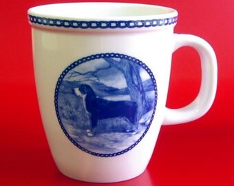 May include: White ceramic mug with a blue and white design of a Bernese Mountain Dog standing in a snowy forest. The mug has a blue rim and a blue handle.