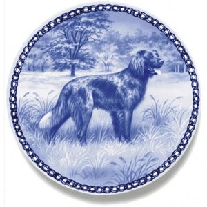 Könnte beinhalten: Ein blauer und weißer Keramikteller mit einem dekorativen Rand in Kettengliedform. Der Teller zeigt einen schwarz-weißen Hund, der in einem Feld mit hohem Gras steht.