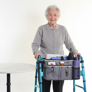 May include: A senior woman stands with a blue walker, which has a gray organizer bag attached. The bag contains various items, including a water bottle, umbrella, and medication. A small, round table is to the left.