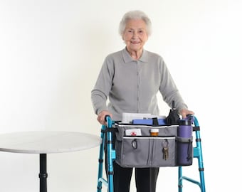 May include: A senior woman stands with a blue walker, which has a gray organizer bag attached. The bag contains various items, including a water bottle, umbrella, and medication. A small, round table is to the left.