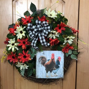 May include: A wreath made of grapevine with red and white flowers, green leaves, and a black and white checkered bow. A sign with two roosters is attached to the wreath.
