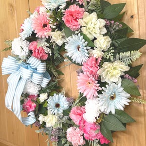 May include: A decorative wreath featuring an assortment of artificial flowers in shades of light blue, soft pink, and cream, accented with green foliage and a large, light blue and white striped fabric bow attached to the left side of the arrangement.