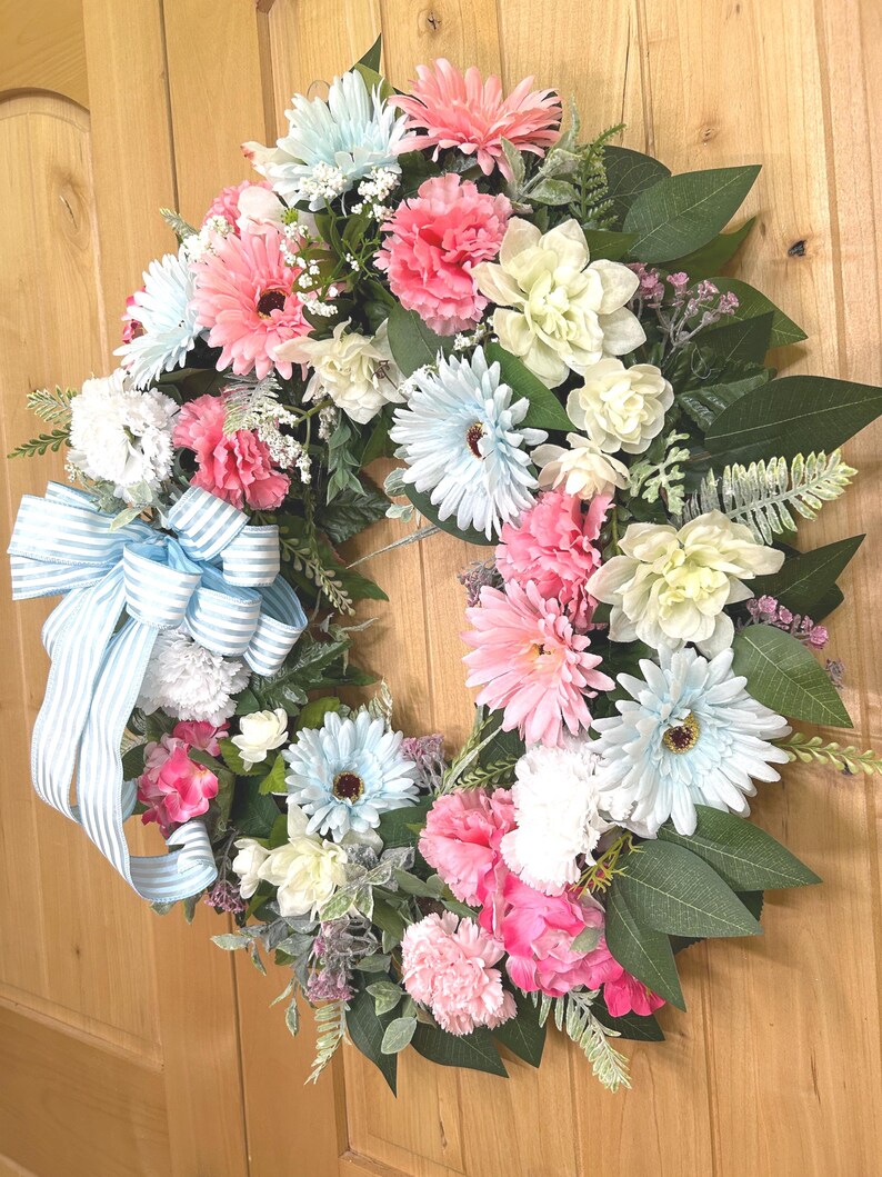 May include: A decorative wreath featuring an assortment of artificial flowers in shades of light blue, soft pink, and cream, accented with green foliage and a large, light blue and white striped fabric bow attached to the left side of the arrangement.