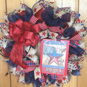 May include: A decorative wreath featuring red, white, and blue mesh ribbons, burlap ribbons with star patterns, and a large red glittery bow. A rectangular sign is attached to the front, displaying a star graphic and the word AMERICA in white.