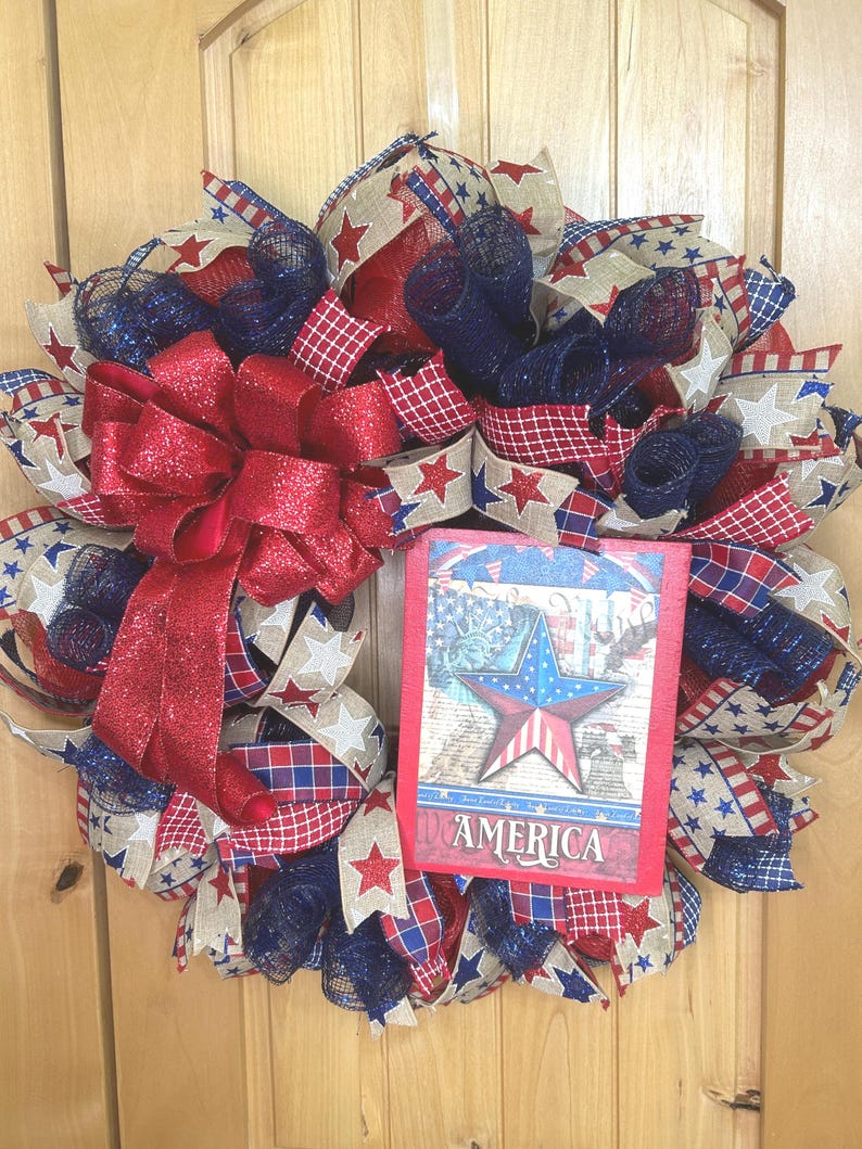 May include: A decorative wreath featuring red, white, and blue mesh ribbons, burlap ribbons with star patterns, and a large red glittery bow. A rectangular sign is attached to the front, displaying a star graphic and the word AMERICA in white.