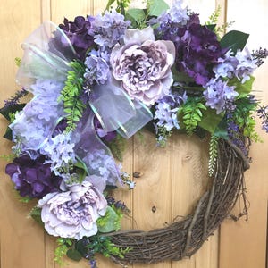 May include: A circular wreath constructed from brown twigs, adorned with a collection of artificial flowers in shades of deep purple, light lavender, and soft mauve. A translucent, iridescent ribbon is tied into a bow among the foliage.