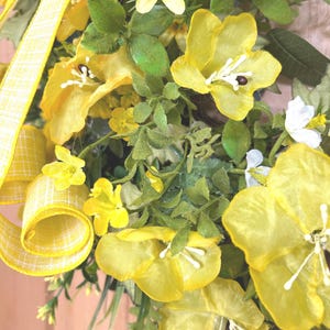 May include: A decorative wreath featuring bright yellow fabric flowers with white stamens, interspersed with green foliage and small white blossoms. A yellow and white checkered ribbon is woven through the arrangement, adding texture.