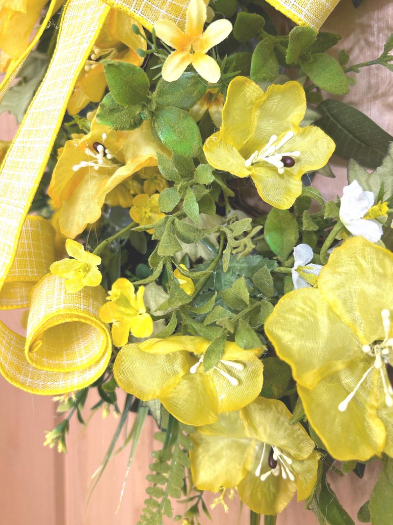May include: A decorative wreath featuring bright yellow fabric flowers with white stamens, interspersed with green foliage and small white blossoms. A yellow and white checkered ribbon is woven through the arrangement, adding texture.