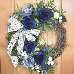 Op de afbeelding: Een ronde krans op een basis van bruine takken, versierd met kunstmatige donkerblauwe en lichtgrijze rozen, kleine witte bloemen en groen blad. Een wit stoffen lint met een blauw bloemenpatroon is aan de linkerkant vastgemaakt.
