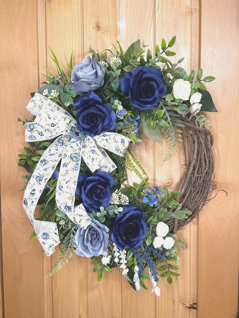 Op de afbeelding: Een ronde krans op een basis van bruine takken, versierd met kunstmatige donkerblauwe en lichtgrijze rozen, kleine witte bloemen en groen blad. Een wit stoffen lint met een blauw bloemenpatroon is aan de linkerkant vastgemaakt.