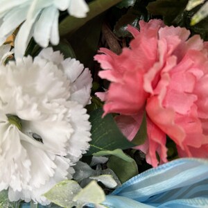 May include: A floral arrangement featuring a large white carnation and a vibrant pink carnation with ruffled petals. The bouquet includes green foliage and a light blue sheer ribbon with thin stripes, all set against a natural brown twig base.