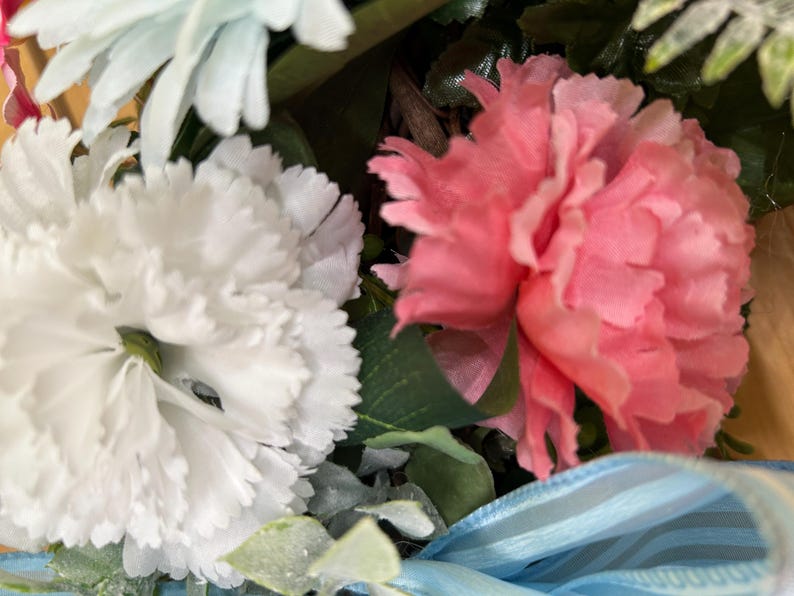 May include: A floral arrangement featuring a large white carnation and a vibrant pink carnation with ruffled petals. The bouquet includes green foliage and a light blue sheer ribbon with thin stripes, all set against a natural brown twig base.