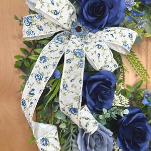 Op de afbeelding: Een decoratieve krans met kunstmatige rozen in diep marineblauw en leigrijs, geaccentueerd met groen blad en kleine witte bloemtakjes. Een cr&egrave;mekleurig jute lint met een blauw rozen- en rankenpatroon is bovenaan tot een strik gebonden.