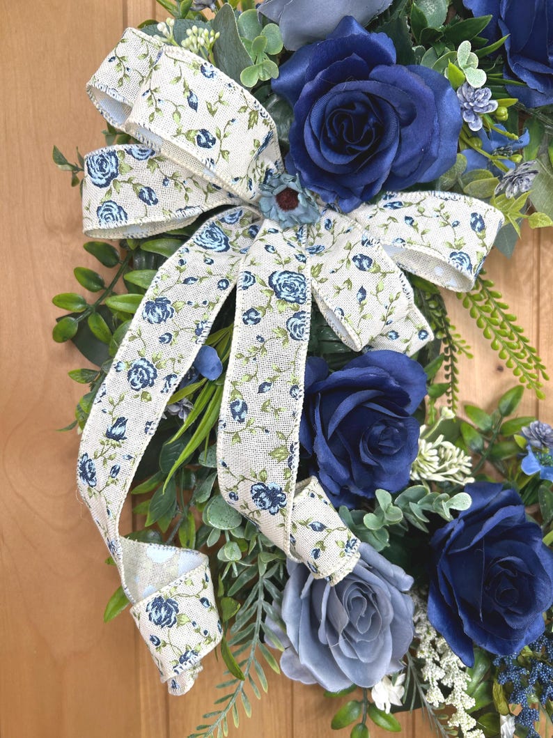 Op de afbeelding: Een decoratieve krans met kunstmatige rozen in diep marineblauw en leigrijs, geaccentueerd met groen blad en kleine witte bloemtakjes. Een cr&egrave;mekleurig jute lint met een blauw rozen- en rankenpatroon is bovenaan tot een strik gebonden.