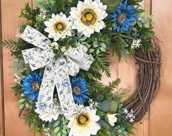 Op de afbeelding: Een ronde krans gemaakt van bruine takken, versierd met witte en blauwe kunstbloemen, weelderig groen blad en varens. Een decoratieve stoffen strik met een blauw bloemenpatroon op een crèmekleurige achtergrond is aan de linkerkant bevestigd.