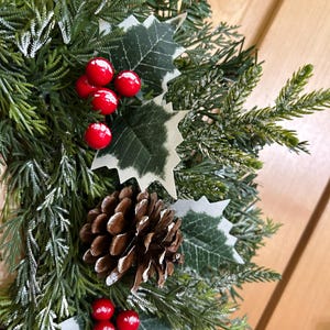 May include: A decorative wreath featuring artificial green pine needles with a light dusting of white snow. The arrangement includes clusters of bright red holly berries, dark green holly leaves with white edges, and a single brown pinecone.