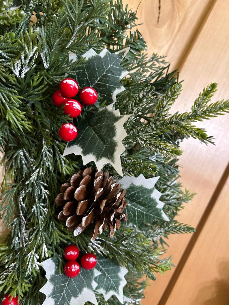 May include: A decorative wreath featuring artificial green pine needles with a light dusting of white snow. The arrangement includes clusters of bright red holly berries, dark green holly leaves with white edges, and a single brown pinecone.