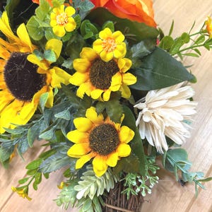 May include: A floral arrangement featuring bright yellow sunflowers, orange roses, and white chrysanthemums accented with green foliage and small yellow blossoms. The flowers are gathered at the base into a bundle of brown, woven twigs.