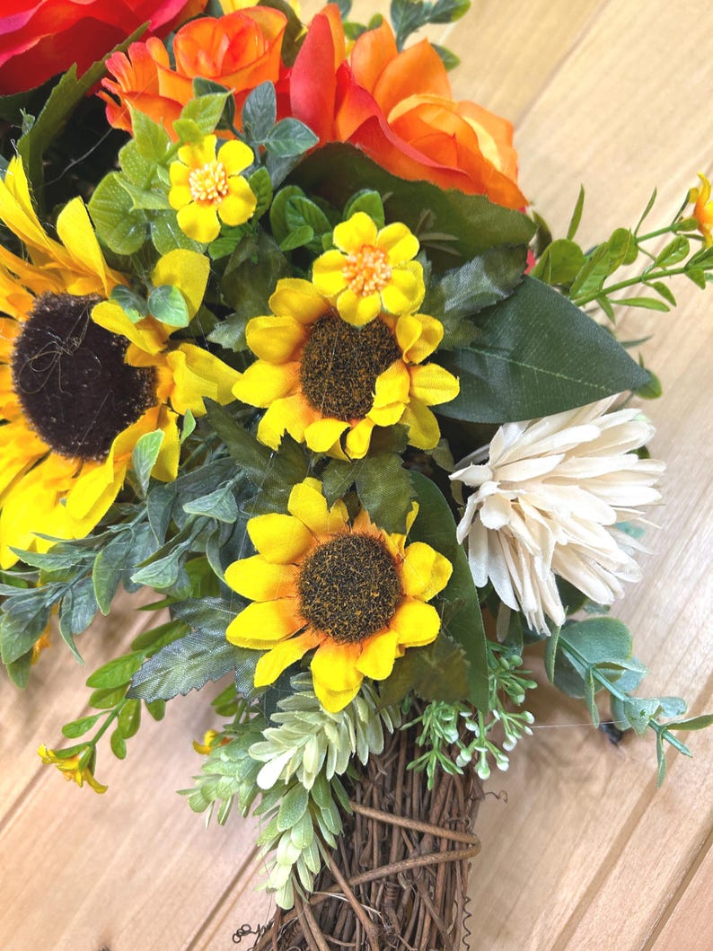 May include: A floral arrangement featuring bright yellow sunflowers, orange roses, and white chrysanthemums accented with green foliage and small yellow blossoms. The flowers are gathered at the base into a bundle of brown, woven twigs.