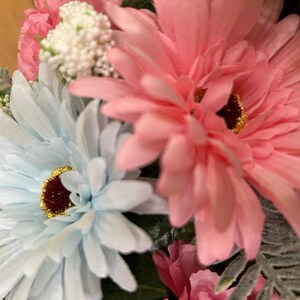 May include: A floral arrangement featuring large, light blue and soft pink daisy-like flowers with dark brown centers and gold glitter accents. Small white clusters of buds, green foliage, and additional pink petals are interspersed throughout.
