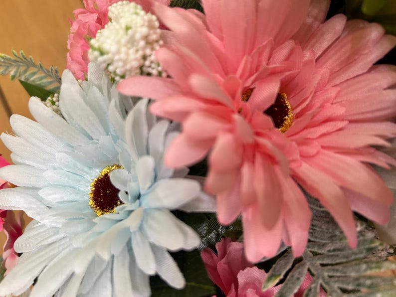 May include: A floral arrangement featuring large, light blue and soft pink daisy-like flowers with dark brown centers and gold glitter accents. Small white clusters of buds, green foliage, and additional pink petals are interspersed throughout.