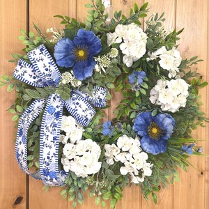 May include: A circular wreath featuring green foliage, white hydrangea clusters, and large blue poppy-like flowers. A decorative ribbon with a blue and white floral pattern and striped edges is tied to the left side, with long tails hanging down.