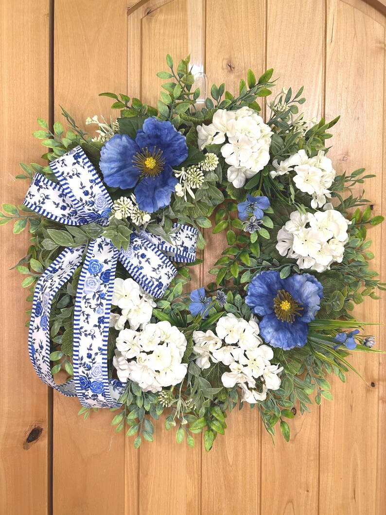 May include: A circular wreath featuring green foliage, white hydrangea clusters, and large blue poppy-like flowers. A decorative ribbon with a blue and white floral pattern and striped edges is tied to the left side, with long tails hanging down.
