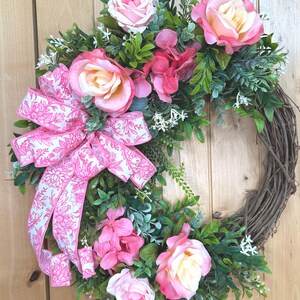 May include: A circular wreath constructed from brown twigs, decorated with artificial pink and cream roses, small green leaves, and white filler flowers. A large, multi-looped fabric bow with a pink floral pattern is attached to the left side.