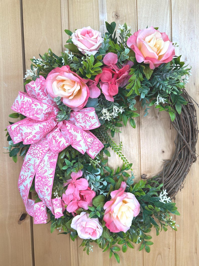 May include: A circular wreath constructed from brown twigs, decorated with artificial pink and cream roses, small green leaves, and white filler flowers. A large, multi-looped fabric bow with a pink floral pattern is attached to the left side.