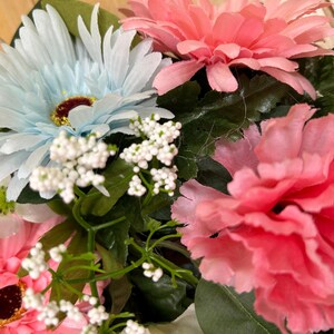 May include: A floral arrangement featuring light blue and pink gerbera daisies, a ruffled pink carnation, and small clusters of white baby's breath flowers. The bouquet includes dark green leaves and stems interspersed among the colorful blossoms.