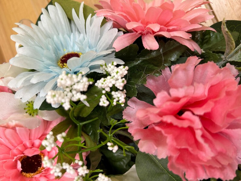 May include: A floral arrangement featuring light blue and pink gerbera daisies, a ruffled pink carnation, and small clusters of white baby's breath flowers. The bouquet includes dark green leaves and stems interspersed among the colorful blossoms.