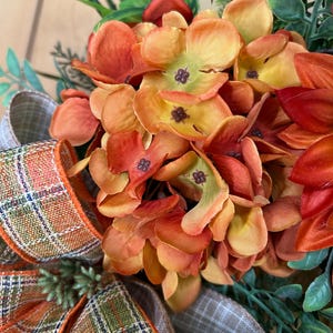 May include: A decorative wreath featuring clusters of orange and yellow silk hydrangea flowers, dark red petals, and green foliage. A wired ribbon with a plaid pattern in orange, green, and white is tied at the base of the floral arrangement.