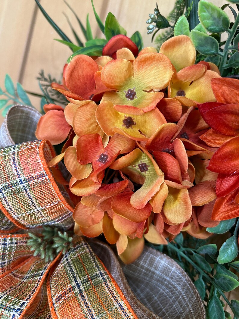 May include: A decorative wreath featuring clusters of orange and yellow silk hydrangea flowers, dark red petals, and green foliage. A wired ribbon with a plaid pattern in orange, green, and white is tied at the base of the floral arrangement.