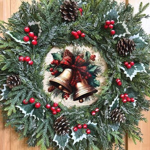 May include: A circular wreath made of green artificial pine branches, dusted with white frost. It features brown pinecones, clusters of bright red berries, and green holly leaves. The center displays an illustration of two gold bells with a red plaid bow.