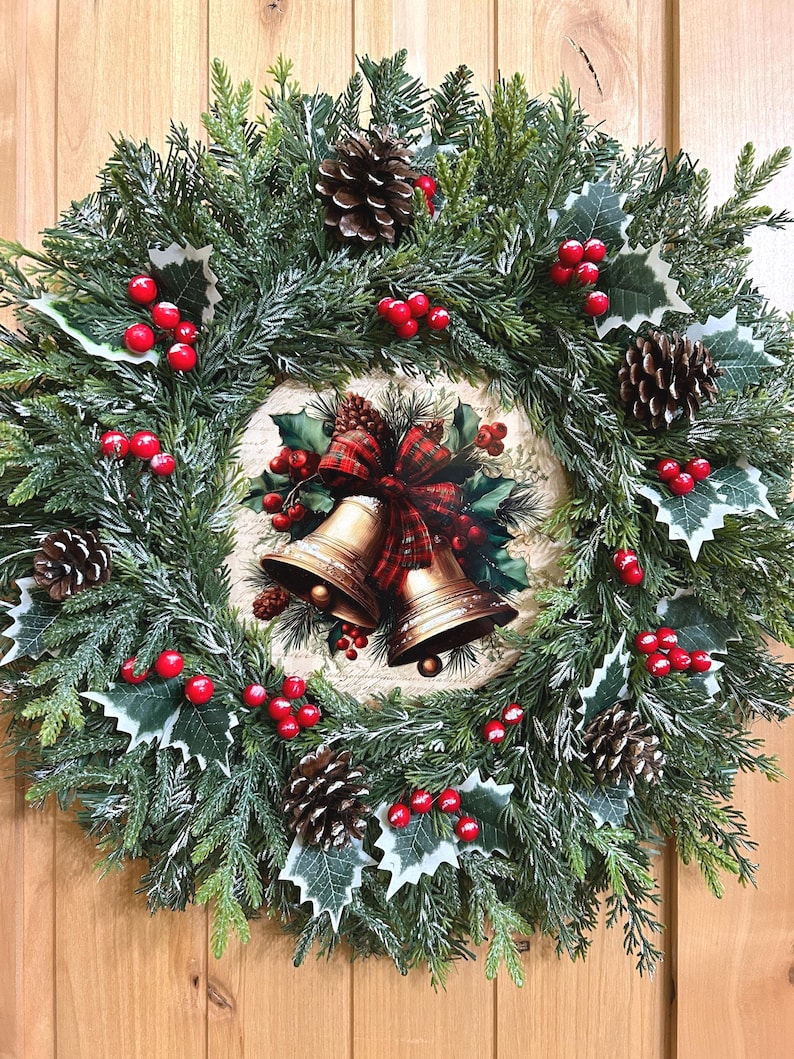 May include: A circular wreath made of green artificial pine branches, dusted with white frost. It features brown pinecones, clusters of bright red berries, and green holly leaves. The center displays an illustration of two gold bells with a red plaid bow.