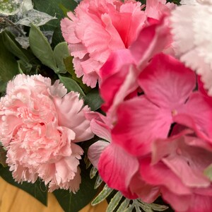 May include: A floral arrangement featuring light pink carnations, vibrant dark pink hydrangea blossoms, and a single light blue flower. The bouquet includes various green leaves and fern-like foliage interspersed among the colorful petals.