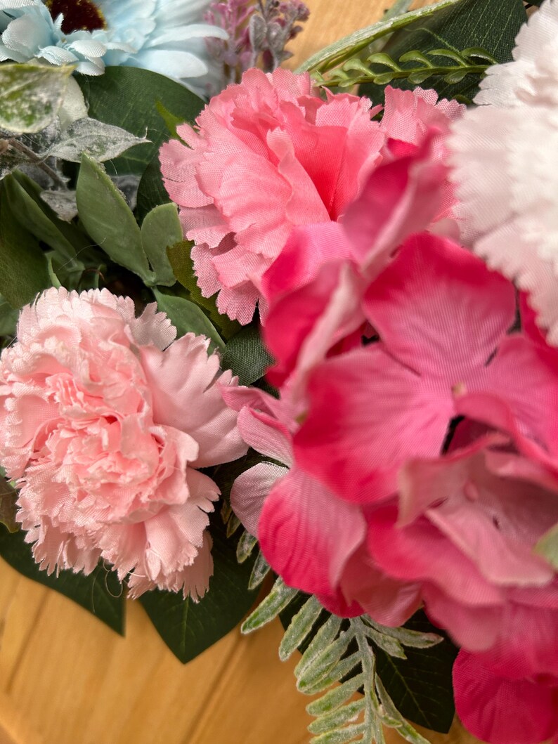 May include: A floral arrangement featuring light pink carnations, vibrant dark pink hydrangea blossoms, and a single light blue flower. The bouquet includes various green leaves and fern-like foliage interspersed among the colorful petals.