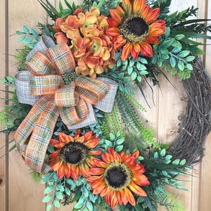 May include: A decorative wreath featuring three large orange and yellow sunflowers with dark brown centers, a cluster of orange hydrangea flowers, and various green foliage. A plaid ribbon in shades of orange, gray, and white is tied to the side.