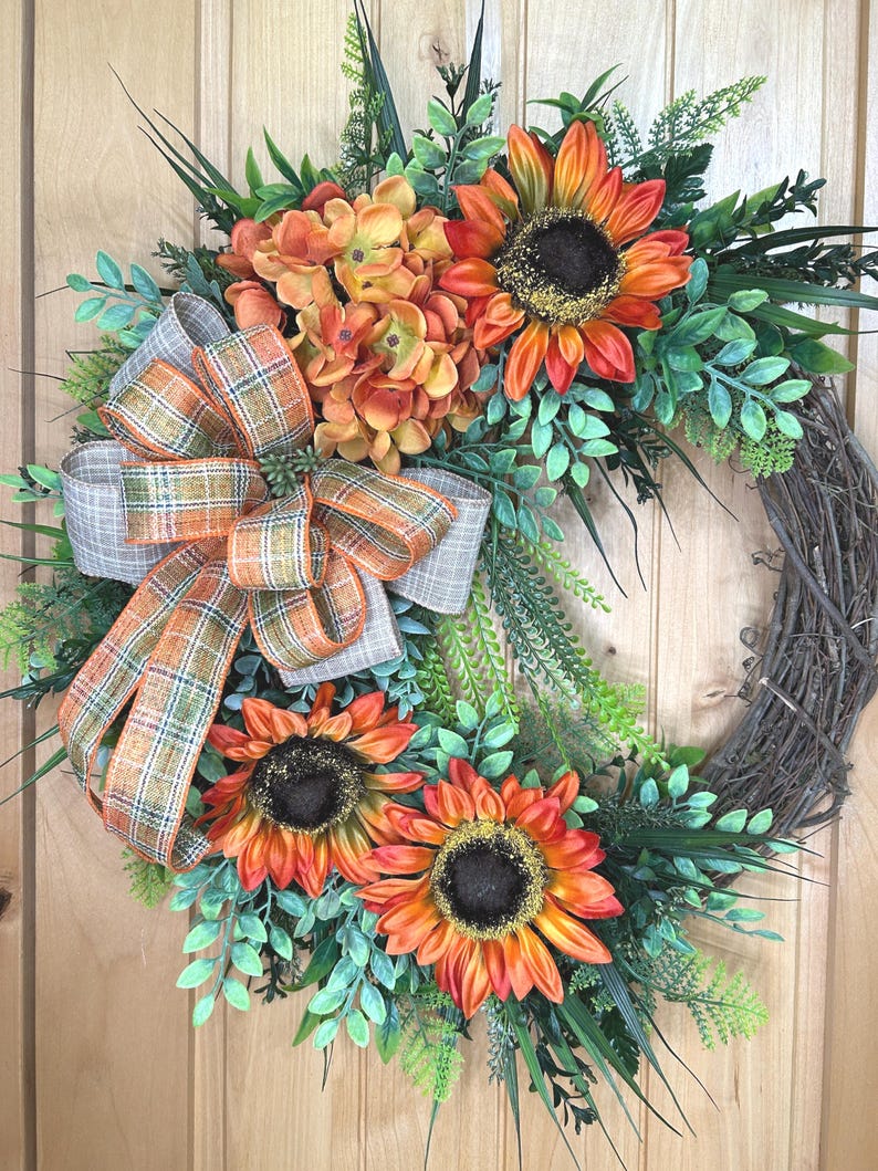 May include: A decorative wreath featuring three large orange and yellow sunflowers with dark brown centers, a cluster of orange hydrangea flowers, and various green foliage. A plaid ribbon in shades of orange, gray, and white is tied to the side.